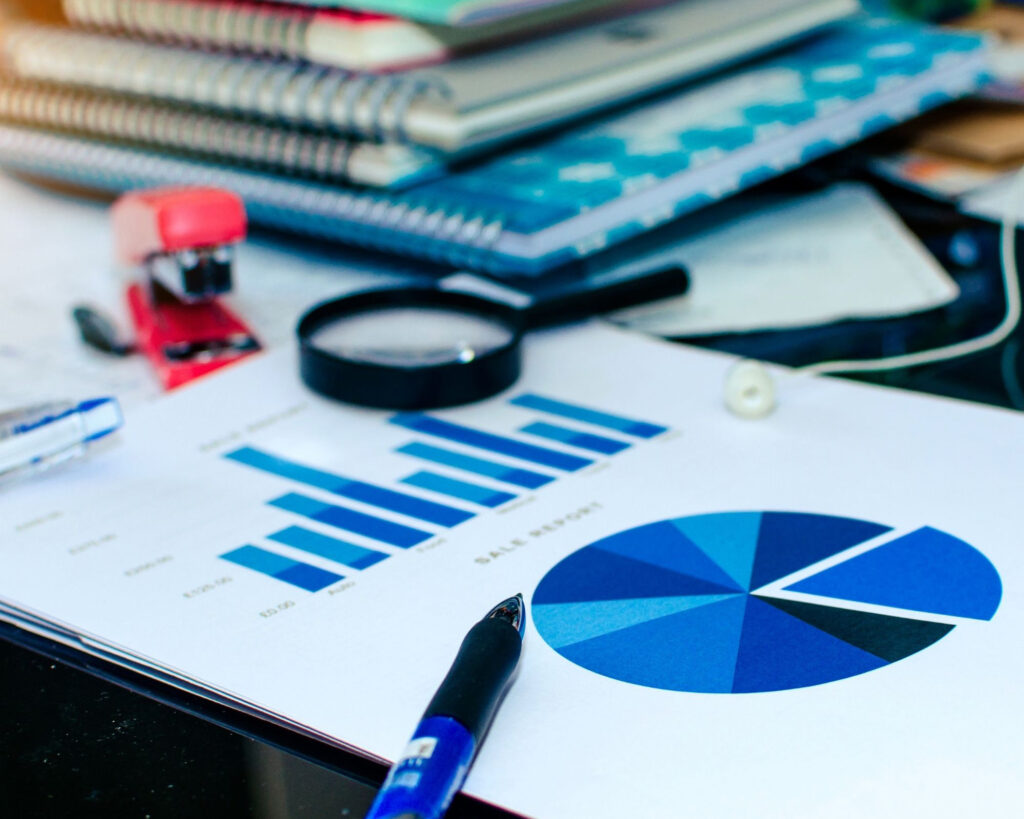 Close-up of business workspace with printed charts, colorful binders, magnifying glass, pen, and office supplies on glass table.