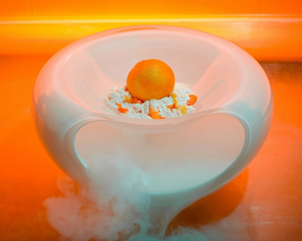 Vibrant orange sphere on swirling dish with billowing smoke, against orange background.