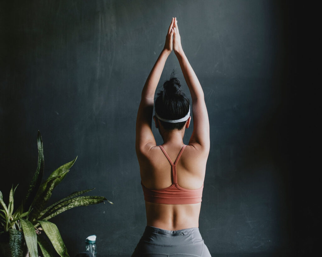 Woman in yoga attire practices seated pose with clasped hands, facing dark wall in peaceful indoor setting.